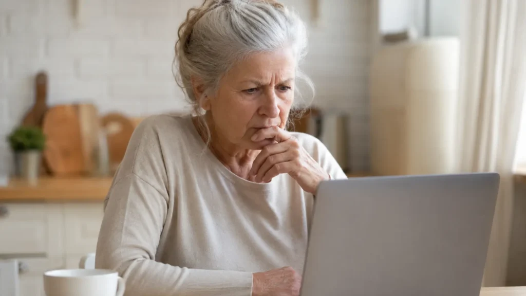 Senior woman with grey hair looking skeptically at a laptop while researching chair yoga evidence, costs, and app reviews for seniors.
