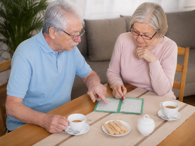 An older couple sitting together at a wooden table sharing tea and pointing at a small puzzle book while conversing.